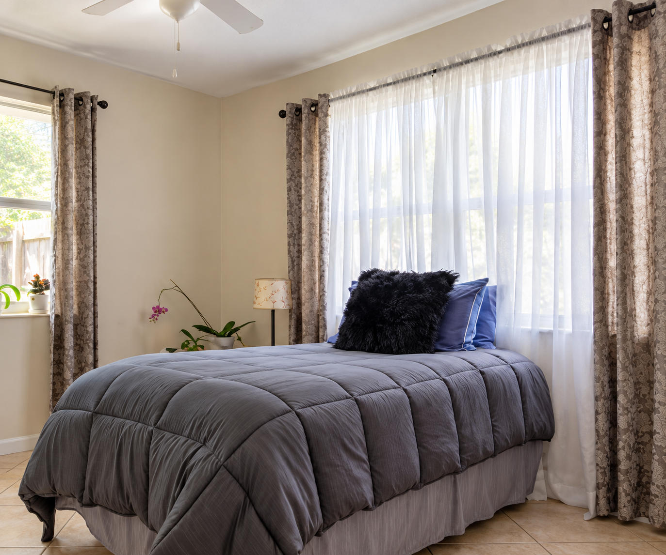 A bright, tidy bedroom with a made bed, sheer curtains, and potted plants